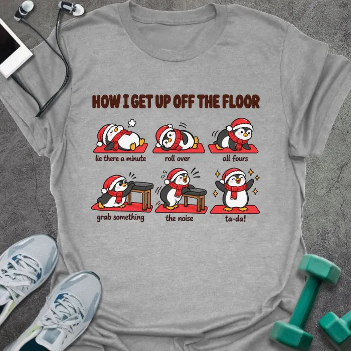 Sport grey t-shirt printed with "HOW I GET UP OFF THE FLOOR" in red, with six penguins in Santa hats showing the steps: lie there a minute, roll over, all fours, grab something, the noise, ta-da!
