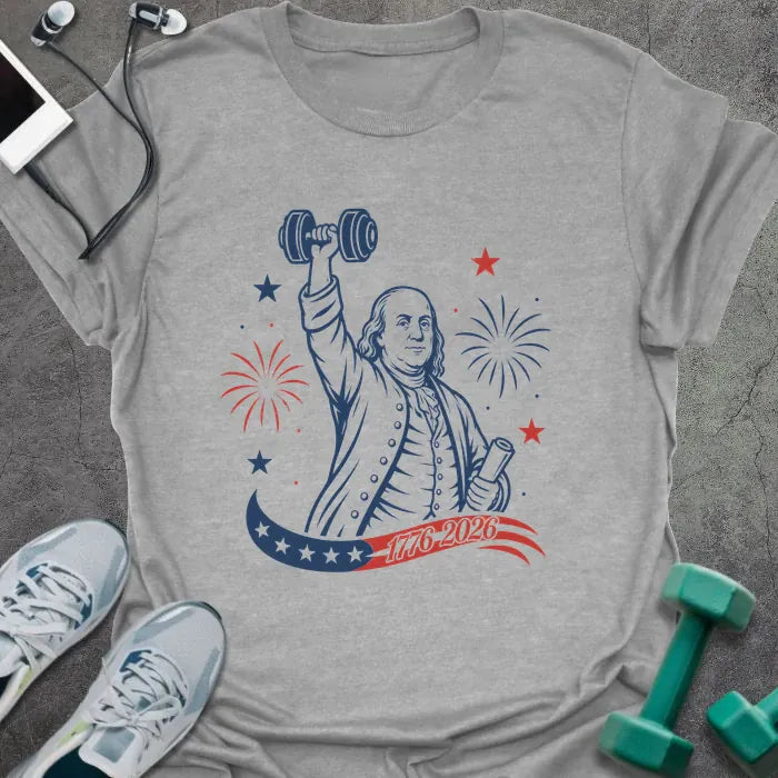 Sport grey t-shirt printed with "1776-2026" in navy, red, with Benjamin Franklin holding a dumbbell overhead and a scroll, fireworks and stars