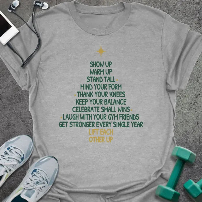 Sport grey t-shirt printed with a Christmas tree made of words, "SHOW UP, WARM UP, STAND TALL, MIND YOUR FORM, THANK YOUR KNEES, KEEP YOUR BALANCE, CELEBRATE SMALL WINS, LAUGH WITH YOUR GYM FRIENDS, GET STRONGER EVERY SINGLE YEAR, LIFT EACH OTHER UP", in green and gold with a gold star on top