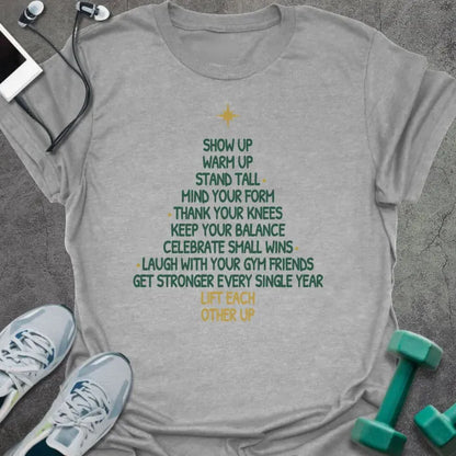 Sport grey t-shirt printed with a Christmas tree made of words, "SHOW UP, WARM UP, STAND TALL, MIND YOUR FORM, THANK YOUR KNEES, KEEP YOUR BALANCE, CELEBRATE SMALL WINS, LAUGH WITH YOUR GYM FRIENDS, GET STRONGER EVERY SINGLE YEAR, LIFT EACH OTHER UP", in green and gold with a gold star on top