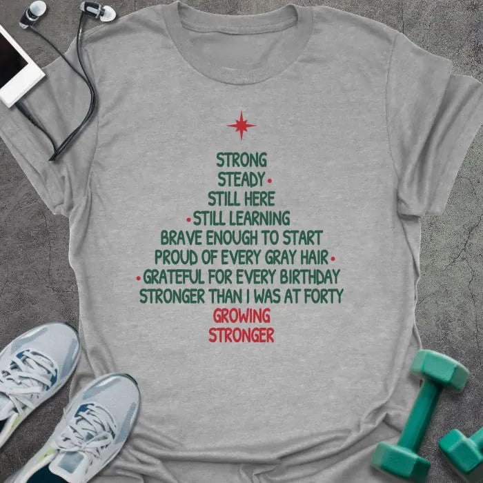 Sport grey t-shirt printed with a Christmas tree made of words, "STRONG, STEADY, STILL HERE, STILL LEARNING, BRAVE ENOUGH TO START, PROUD OF EVERY GRAY HAIR, GRATEFUL FOR EVERY BIRTHDAY, STRONGER THAN I WAS AT FORTY, GROWING STRONGER", in green and red with a red star on top