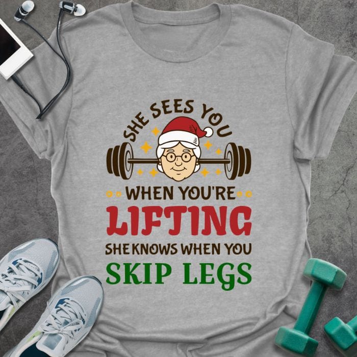 Sport grey t-shirt printed with "She Sees You When You're Lifting Sheknows When You Skip Legs" in black, red, and gold, with female Santa and barbell