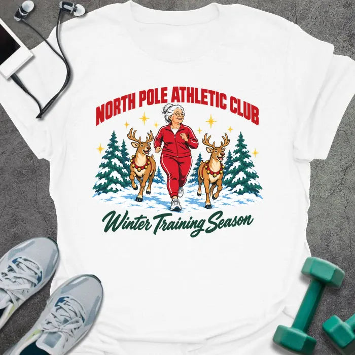 White t-shirt printed with "NORTH POLE ATHLETIC CLUB" in red and "Winter Training Season" in green script, with a white-haired woman in a red tracksuit jogging between two reindeer through snowy pine trees