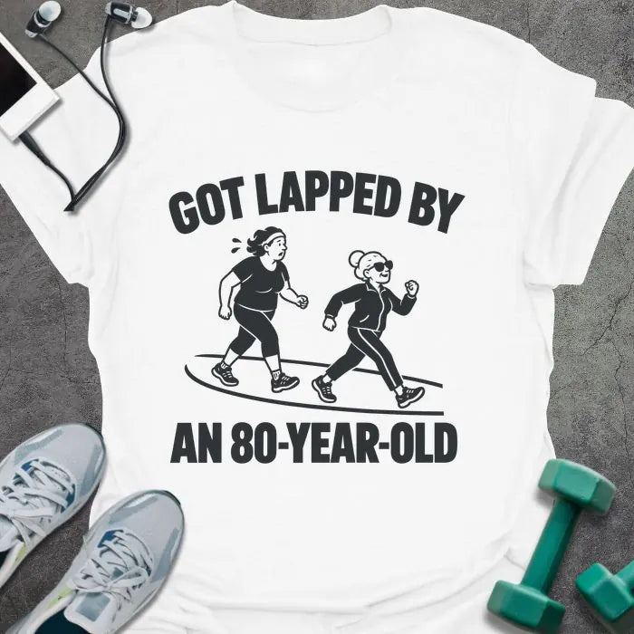 White t-shirt printed with "GOT LAPPED BY AN 80-YEAR-OLD" in black, with a tired jogger being passed by an older woman in sunglasses speed walking on a track