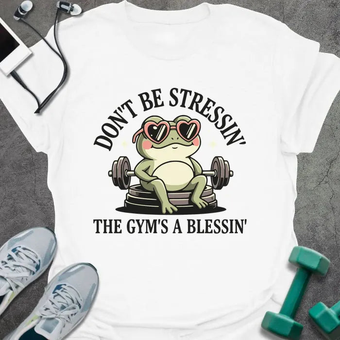 White t-shirt printed with "Don't Be Stressin' the Gym's a Blessin'" in red, with an illustration of a frog with heart-shaped sunglasses lifting dumbbells