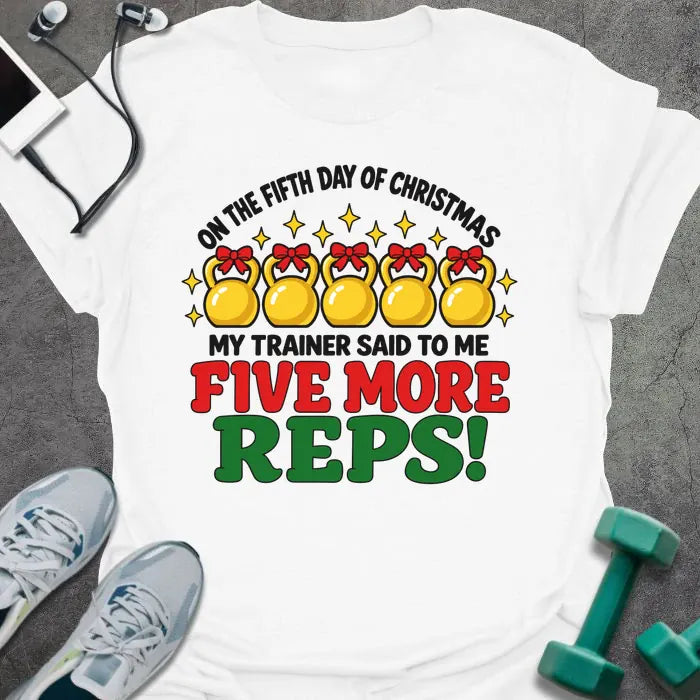 White t-shirt printed with "ON THE FIFTH DAY OF CHRISTMAS MY TRAINER SAID TO ME, FIVE MORE REPS!" in black, red and green, with five gold kettlebells tied with red bows