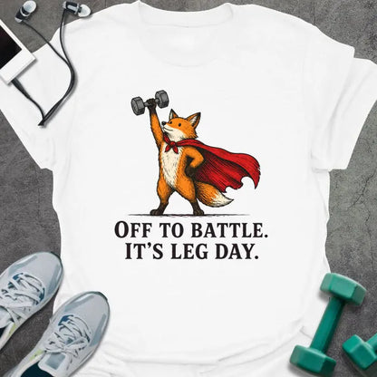 White t-shirt printed with "OFF TO BATTLE. IT'S LEG DAY." in black lettering, with a red fox wearing a red cape and holding a dumbbell