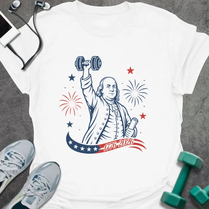 White t-shirt printed with "1776-2026" in navy, red, with Benjamin Franklin holding a dumbbell overhead and a scroll, fireworks and stars