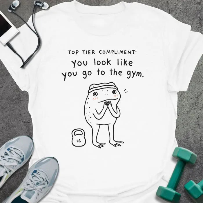 White t-shirt printed with "TOP TIER COMPLIMENT: you look like you go to the gym." in black, with frog graphic holding barbells