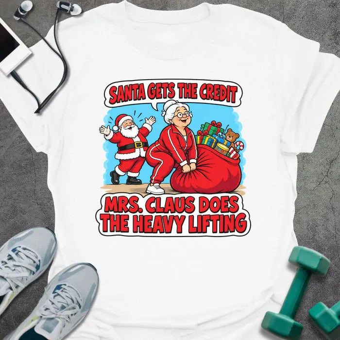 White t-shirt printed with "SANTA GETS THE CREDIT, MRS. CLAUS DOES THE HEAVY LIFTING" in red and white, with Mrs. Claus hauling a sack of gifts while Santa waves