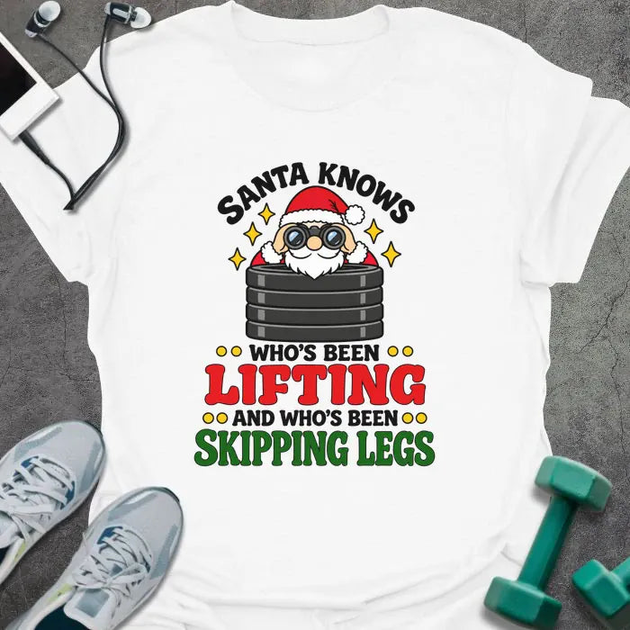 White t-shirt printed with "SANTA KNOWS WHO'S BEEN LIFTING AND WHO'S BEEN SKIPPING LEGS" in black, red and green, with Santa peering through binoculars over a stack of weight plates