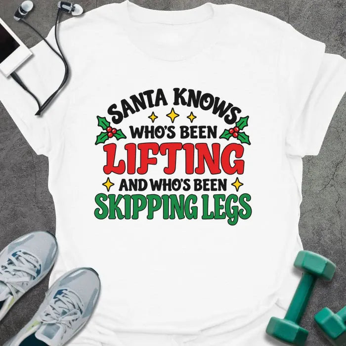 White t-shirt printed with "SANTA KNOWS WHO'S BEEN LIFTING AND WHO'S BEEN SKIPPING LEGS" in black, red and green, with holly and gold stars