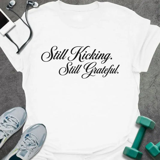 White t-shirt printed with "Still Kicking. Still Grateful." in black italic script