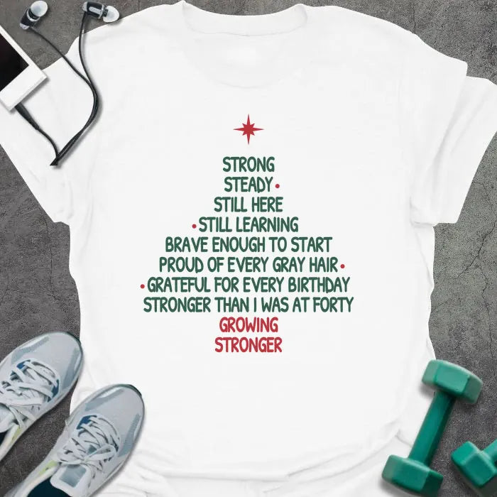 White t-shirt printed with a Christmas tree made of words, "STRONG, STEADY, STILL HERE, STILL LEARNING, BRAVE ENOUGH TO START, PROUD OF EVERY GRAY HAIR, GRATEFUL FOR EVERY BIRTHDAY, STRONGER THAN I WAS AT FORTY, GROWING STRONGER", in green and red with a red star on top