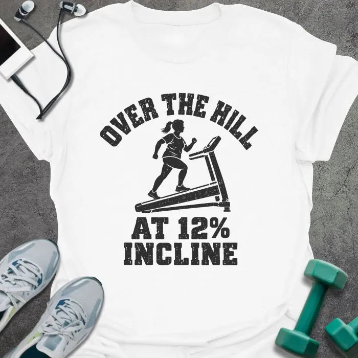 White t-shirt printed with "OVER THE HILL AT 12% INCLINE" in charcoal, with a woman walking on a treadmill