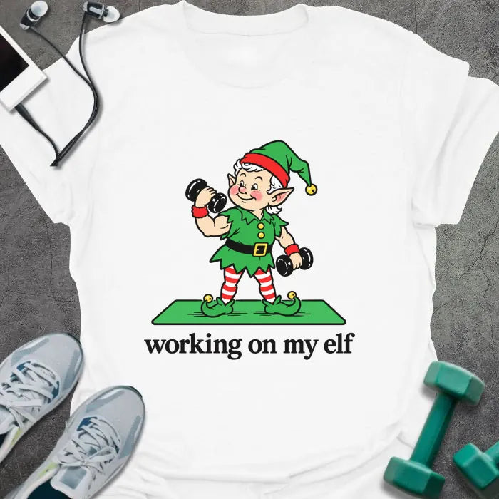 White t-shirt printed with "working on my elf" in black, with a vintage elf lifting dumbbells on a green mat