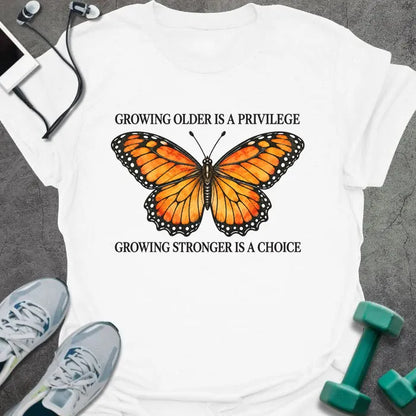White t-shirt printed with "GROWING OLDER IS A PRIVILEGE, GROWING STRONGER IS A CHOICE" in black, with an orange watercolor monarch butterfly