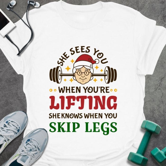 White t-shirt printed with "She Sees You When You're Lifting Sheknows When You Skip Legs" in black, red, and gold, with female Santa and barbell