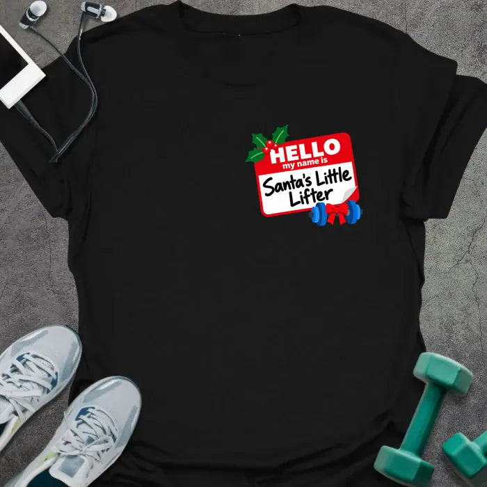 Black t-shirt printed with a small red name tag on the chest reading "HELLO my name is Santa's Little Lifter", with holly and a blue dumbbell tied with a red bow